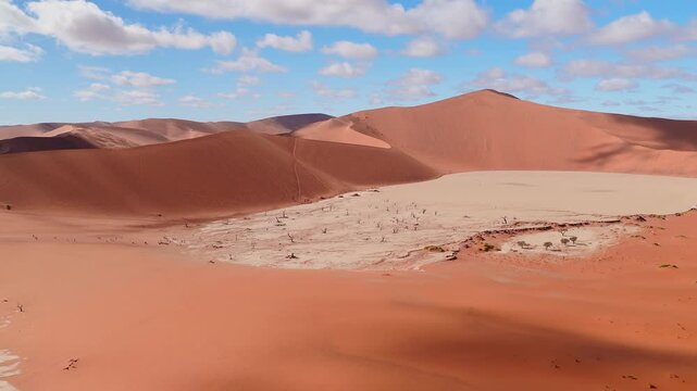Aerial view passes over Sossusvlei dunes to Deadvlei in Namibia, with black camelthorn trees, climbing tracks, tiny people on ridges, cracked pan, and cumulus clouds.