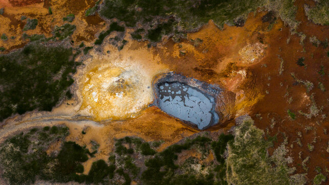 Aerial view of a geothermal mud pool surrounded by vibrant orange and yellow mineral deposits and patches of green moss in the volcanic landscape of Husavik, Nordurthing, Iceland.