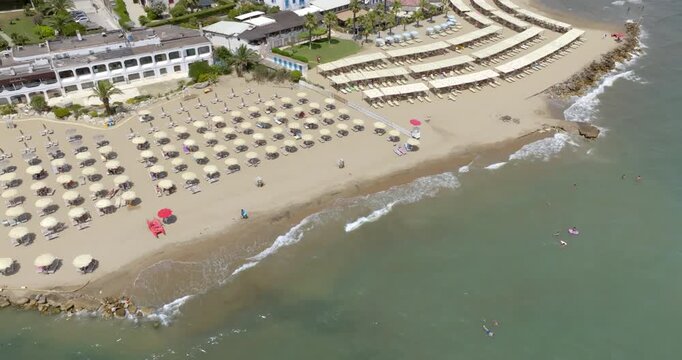 Aerial view of a sandy beach resort lined with neatly arranged rows of sun umbrellas and loungers along gentle turquoise ocean waves, near coastal buildings on a sunny summer day. It's Circeo in Italy