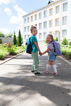 Two smiling elementary school children with backpacks holding hands while walking toward the school building on a sunny morning