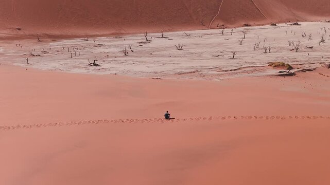Aerial view of Deadvlei in Sossusvlei, Namib Desert, Namibia, with rust red dunes, dead camel thorn trees, a lone person on a ridge, and tiny figures near the pan.
