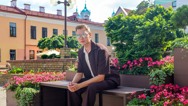 A young handsome guy brown clothes sits on a wooden bench in a busy city square.