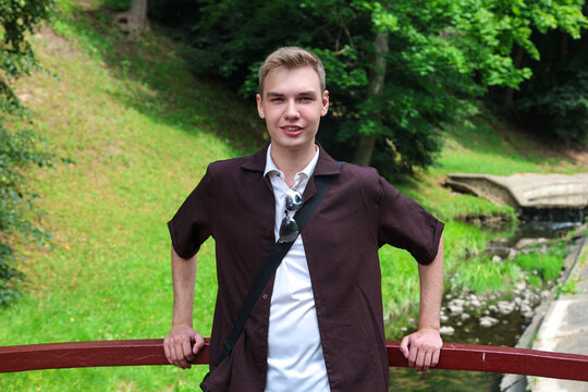 young blond guy in brown clothes stands on a city bridge over a river.