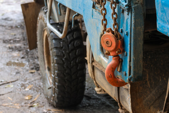 Old industrial tow hook hanging from weathered blue truck with mud covered tire and chain, gritty worksite atmosphere conveying rugged utility and worn texture
