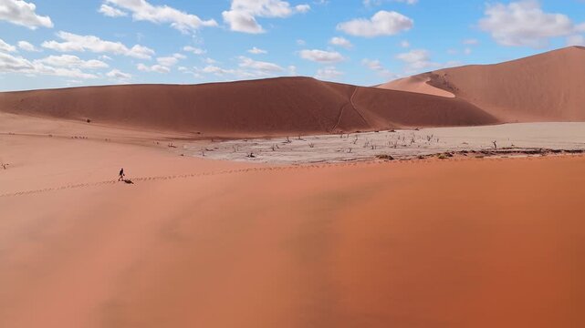 Aerial view of Deadvlei in Namibia shows rust red dunes, white clay pan, dead camelthorn trees, lone person walking with footprints, low sun, blue sky, scattered clouds.