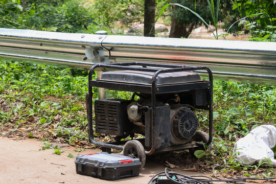 Portable generator on grassy roadside with repair toolbox and cables, rugged metal frame and wheels, outdoor construction equipment providing emergency power for worksite or camping