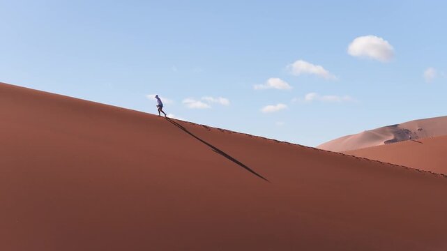 Aerial view shows a lone hiker walking a sharp Sossusvlei dune ridge in the Namib Desert, Namibia. Rust dunes, footprints, pale sky, low sun, long shadows.