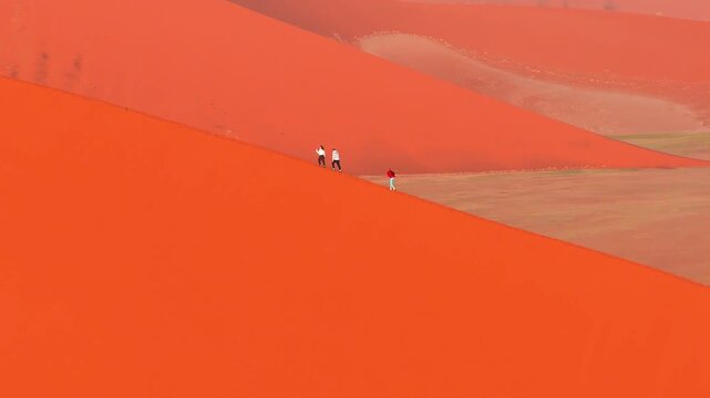 Aerial view of Namib Desert dunes at Sossusvlei, Namibia, as three hikers walk a sharp crest, warm light shapes sweeping sand and pale pan while the camera glides