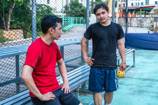 Two male Thai sepak takraw players standing by the sideline of an outdoor court, holding a traditional rattan ball while engaging in a serious tactical discussion during a match pause