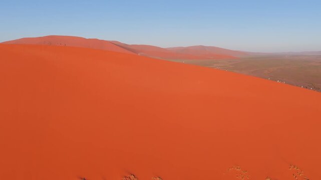 Aerial view of a towering orange red dune in Sossusvlei, Namibia. Small groups walk the knife edge crest as a sweeping low shadow moves in warm morning or late day light.
