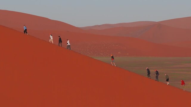 Aerial view shows hikers moving single file on Dune 45 in the Namib Desert near Sesriem, with long shadows, footprints, and soft light across vast red dunes.