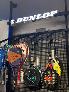 A display of Dunlop sports equipment features several padel rackets in various colors hanging on a grid. The rackets show different designs, showcasing the brand.