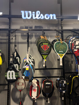 A display of various tennis rackets on a store shelf. Different styles and colors are visible, including branded Wilson rackets. The setting is a sports equipment store.