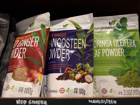Three packets of herbal powders are displayed on a shelf. The pouches include ginger powder, mangosteen powder, and moringa leaf powder. Each package features distinct colors and branding.