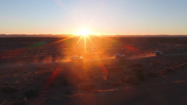 Aerial view of three white SUVs drive in staggered formation across Namibia near Sossusvlei, dust trails rise, low ridge on horizon, golden hour sun and lens flares.