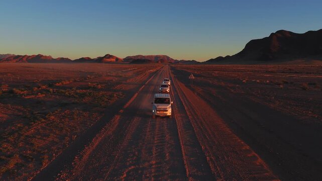 Aerial view shows white vans driving on a gravel road in Namibia toward the Spitzkoppe massif, dust rising in golden hour light with long shadows in the Namib Desert.