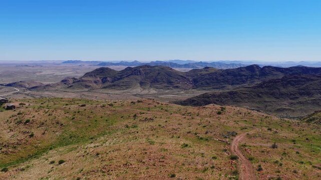 Aerial view over Namibia Damaraland shows rocky hills, broad plains, and a meandering dirt track at midday. Layered ranges recede, sparse shrubs and ochre grasses show depth.