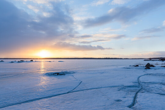 A tranquil frozen lake at sunset with cracked ice patterns leading to a distant shoreline, soft pastel sky and warm sunlight reflecting on the icy surface, evoking calm, solitude and cold beauty