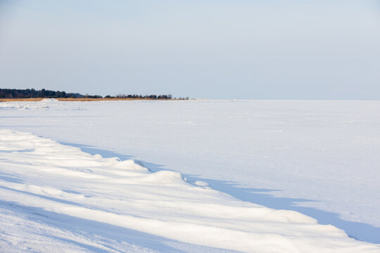 Wide, minimal winter landscape of a snow-covered frozen beach with low dunes and distant trees on the horizon, conveying calm, cold solitude and quiet coastal beauty on a pale morning