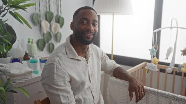 Man resting hand on crib rail in building nursery, seated beside wooden crib, wearing linen shirt and smiling with wedding ring visible to camera; parental pride bonding.