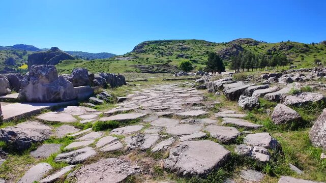 An ancient stone-paved processional road stretches toward rolling green hills under a clear blue sky at Hattusa, Turkey.