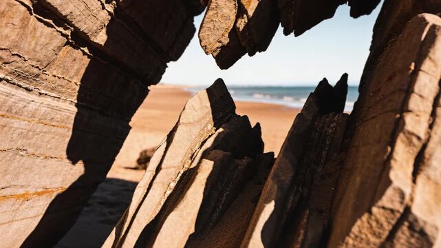 Pushing camera, revealing jagged layered rock shifting to frame sandy beach and distant surf