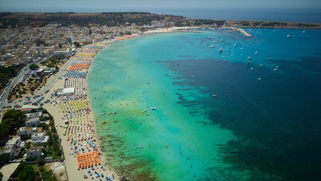 San vito lo capo beach displaying colorful umbrellas and numerous people enjoying the clear turquoise water. With boats dotting the deep blue sea near the coastline and town in trapani. Sicily. Italy