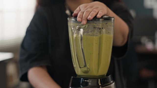 Young adult woman blending green smoothie in kitchen, holding lid of blender. Tilt up close up shot ideal for healthy lifestyle commercials and wellness marketing campaigns