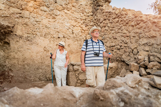 Senior couple tourists with hiking poles and hats exploring ancient stone ruins on summer vacation