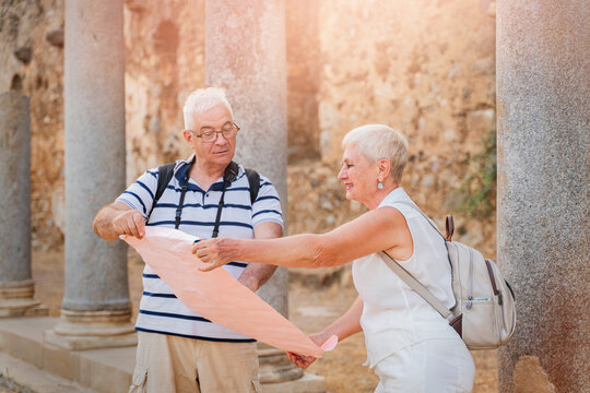 Senior couple tourists reading paper map at ancient Roman ruins with stone columns on summer vacation