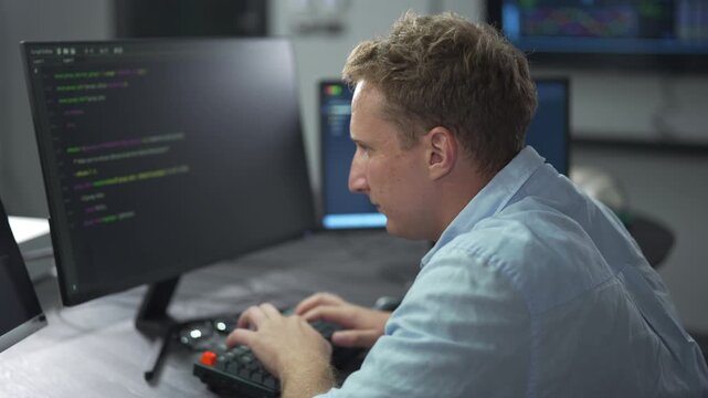 Male software engineer writing program code on multiple desktop screens in a modern office.