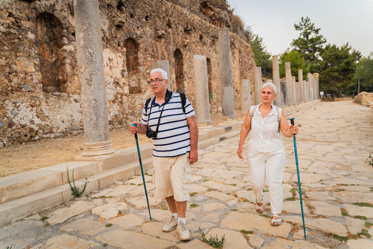 Senior couple tourists with hiking poles and hats exploring ancient stone ruins on summer vacation