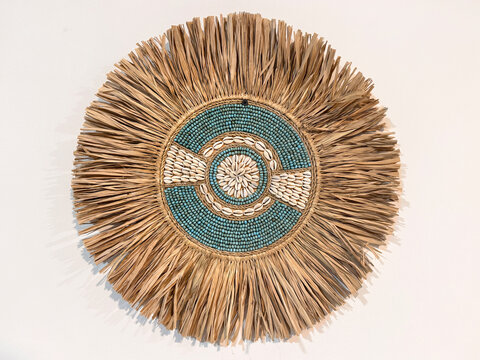 Close up of bohemian circular wall decor featuring natural raffia fringe surrounding turquoise beads and cowrie shells against a white wall. White isolated background