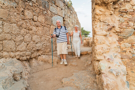 Senior couple tourists with hiking poles and hats exploring ancient stone ruins on summer vacation