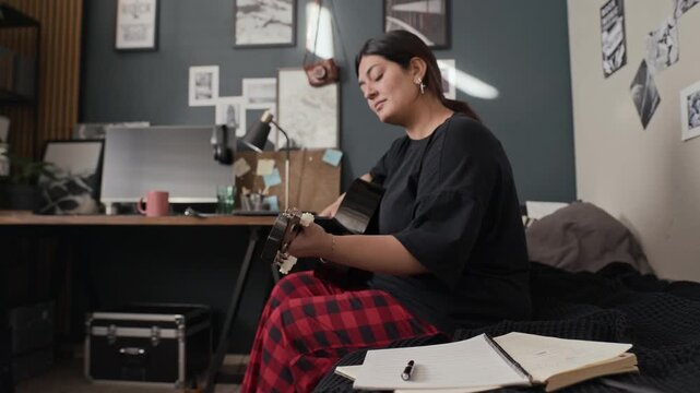Young adult woman writing in notebook and playing acoustic guitar on bed, camera tilting up. Video is ideal for ads promoting online music lessons or creative hobbies