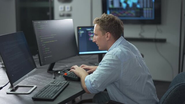 Male software engineer writing program code on multiple desktop screens in a modern office.