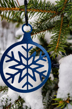 A snowflake ornament is suspended from an evergreen tree branch covered with white snow. The scene shows a natural winter landscape during the holiday time