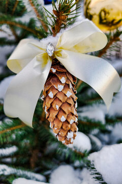 A Christmas tree stands in the snow with a pinecone ornament tied with a ribbon. The scene shows winter in a joyful holiday setting