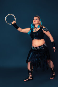 Curvy woman performs with tambourine. Female dancer attired in black ensemble that has been crafted by hand. Outfit consists of leather top and skirt, in addition to pair of bracers, gladiator boots