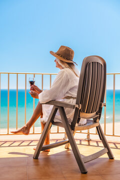 Woman relaxing with glass of red wine on seaside balcony chair enjoying peaceful summer vacation by turquoise ocean