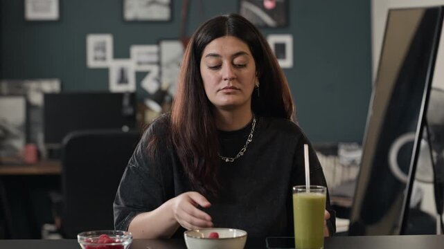 Young adult woman putting fresh raspberries in bowl, scrolling on smartphone and drinking green smoothie. Perfect for wellness apps, healthy lifestyle blogs and nutrition marketing