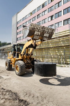A wheel loader lifts and relocates large concrete sewer rings at a construction site. The work is taking place in an urban setting with buildings nearby