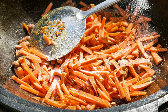 Sliced carrots are being cooked in a kazan with spices added. The preparation is for pilaf, a dish typical in many cultures