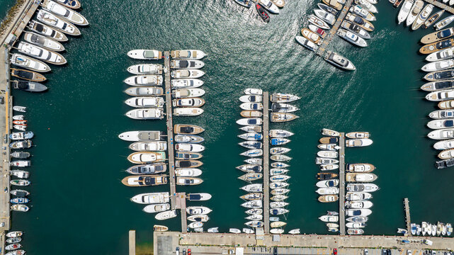 Overhead top-down view of a bustling marina showcasing rows of luxury yachts, motorboats, and sailing vessels docked along floating piers in deep turquoise sea water. Zenithal shot.