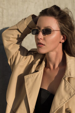 trendy portrait of a stylish woman in a beige trench coat and small Y2K sunglasses under hard sunlight. Pretty hot beauty lady at sunset