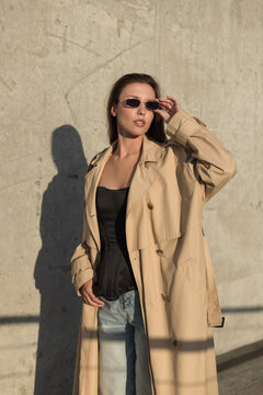 Full-length shot of a stylish woman adjusting her small sunglasses, wearing a beige trench coat and black corset top against a concrete wall in warm golden sunlight. Trendy urban fashion.