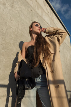 A low-angle shot of a stylish woman in an oversized beige trench coat, black corset, and jeans. She adjusts her sunglasses, holding a large black bag against a concrete wall with dramatic shadows.