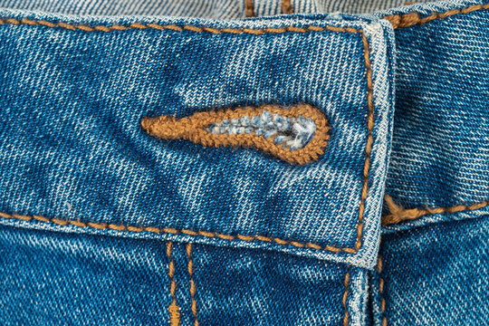 Close-up of denim fabric with buttonhole. Macro detail of blue jeans waistband, stitching, and textile texture. Fashion industry garment production and vintage clothing background concept
