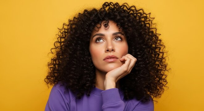 Thoughtful Young Woman with Voluminous Curly Hair Looking Up Against Bright Yellow Background