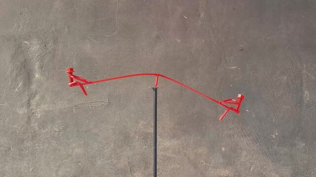 An artistic miniature seesaw, meticulously crafted from bent metal wire and bolts, and painted a vibrant red. The two seats are ingenious representations of small human figures, complete with bolt hea